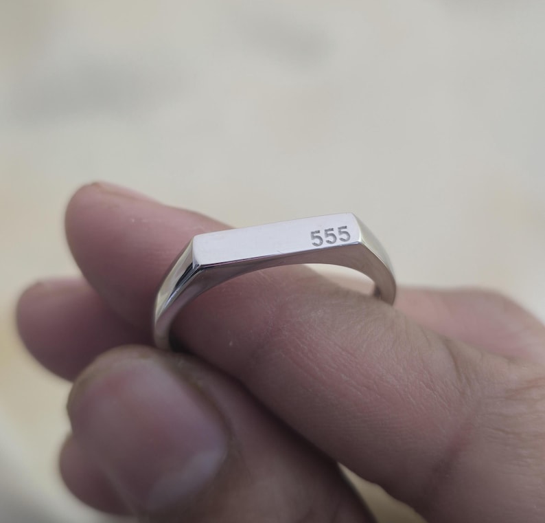 Puede incluir: Un anillo de sello plateado con el n&uacute;mero "555" grabado en la cara rectangular. El anillo est&aacute; sostenido por los dedos de una persona, mostrando su superficie pulida y reflectante. El anillo es una pieza de joyer&iacute;a simple y elegante.