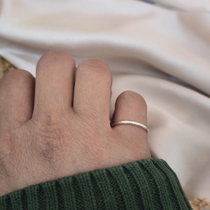 Puede incluir: Un anillo fino de plata que se lleva en un dedo. El anillo tiene un dise&ntilde;o de banda simple, con una superficie lisa y pulida. El dedo est&aacute; contra un fondo suave de tonos neutros, y la persona lleva un su&eacute;ter verde.