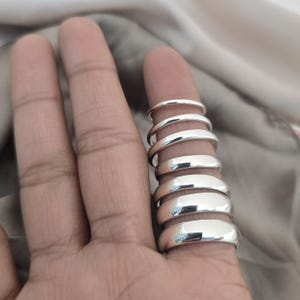 Puede incluir: Una colecci&oacute;n de anillos de plata pulida de diferentes anchos apilados en un dedo. Los anillos tienen una superficie lisa y reflectante, y la luz atrapa sus bordes redondeados. Los anillos se muestran sobre un fondo neutro.