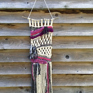 May include: A woven wall hanging with a natural wood dowel at the top. The hanging features a variety of textures and colors, including white, black, and pink.