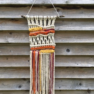 Puede incluir: Tapiz de pared de macramé hecho a mano con una rama de madera natural y cordón de algodón color crema. El diseño incorpora secciones tejidas con acentos de hilo naranja, amarillo y burdeos, y una larga franja color crema.