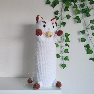 May include: White crocheted cat plush toy with brown ears and paws. The cat is wearing a pink collar with a yellow pacifier. The toy is sitting on a white shelf in front of a green ivy plant.
