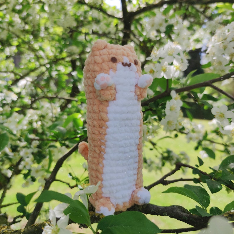 May include: A crocheted plush toy shaped like a cat, with a peach and white color scheme. The toy is sitting on a branch of a tree with white blossoms.