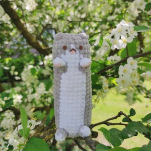 May include: A gray and white crocheted stuffed animal shaped like a cat with a long body. The cat is sitting on a branch of a tree with white flowers.