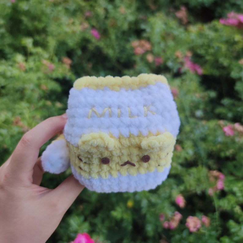 May include: A crocheted milk carton plush toy with a yellow and white striped design. The carton has a cute cartoon face and the word "MILK" embroidered on the front.