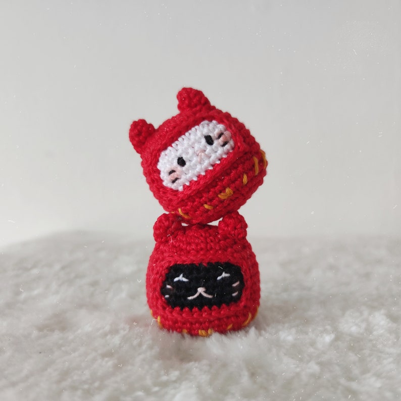 May include: Two crocheted cat toys stacked on top of each other. The top cat is red with white eyes and a yellow mouth. The bottom cat is red with black eyes and a yellow mouth.