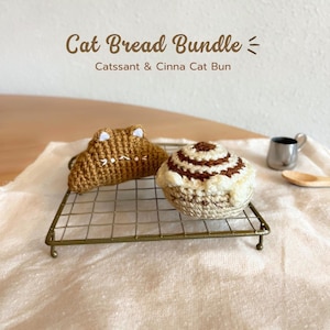 Könnte beinhalten: Zwei gehäkelte katzenförmige Brote auf einem Drahtgestell. Das Katzenbrot links ist braun und croissantförmig. Das Katzenbrot rechts ist weiß mit braunen Wirbeln und cinnamon bun-förmig. Der Text "Cat Bread Bundle" und "Catssant & Cinna Cat Bun" befindet sich über dem Katzenbrot.