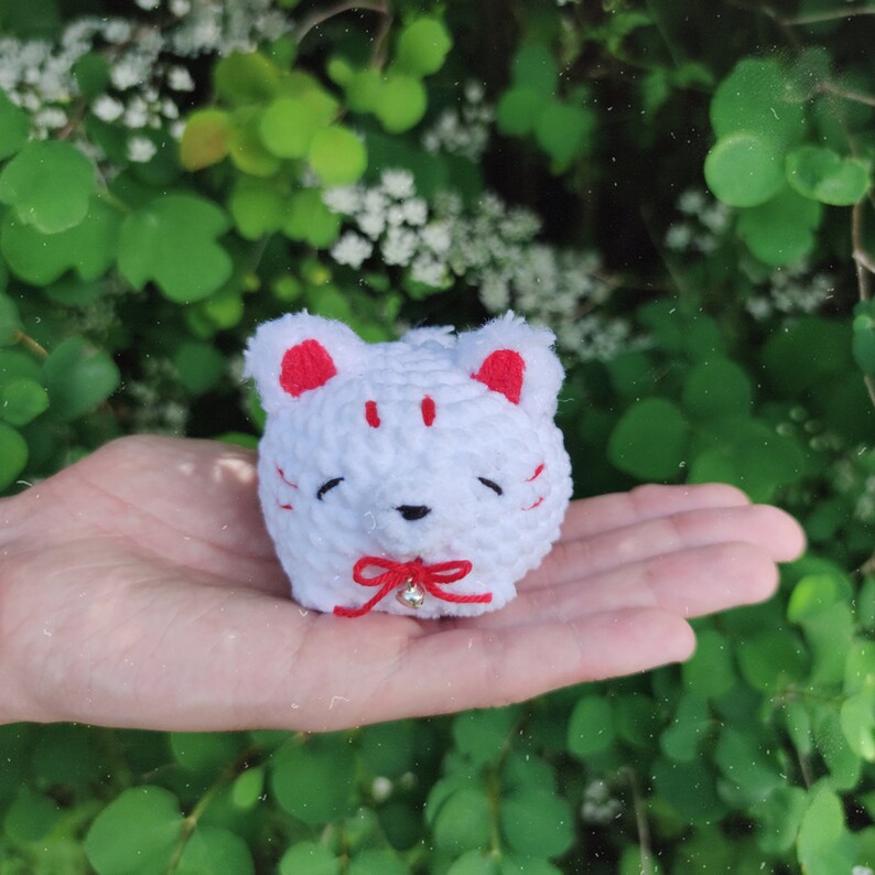 May include: White crocheted cat plushie with red ears and a red bow. The plushie has a small bell hanging from the bow.