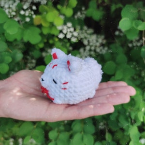 May include: White crocheted fox plushie with red accents. The fox is sitting in a hand.