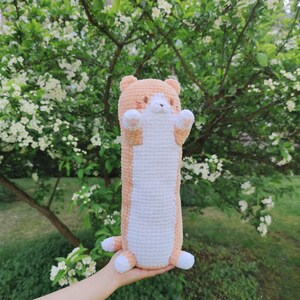 May include: A crocheted stuffed animal shaped like a cat with orange and white fur. The cat is holding its paws up in the air and has a cute expression on its face.