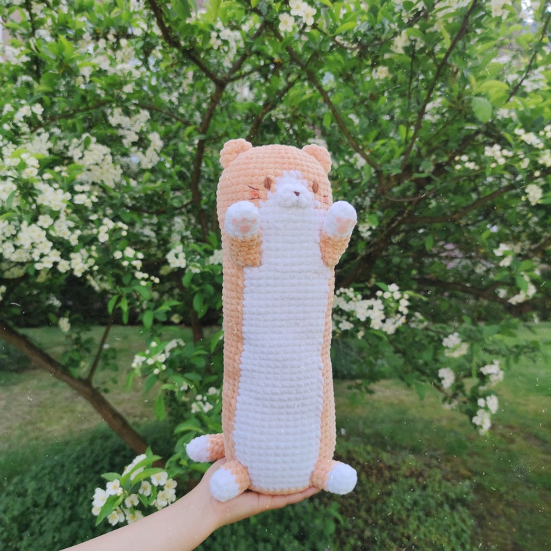 May include: A crocheted stuffed animal shaped like a cat with orange and white fur. The cat is holding its paws up in the air and has a cute expression on its face.