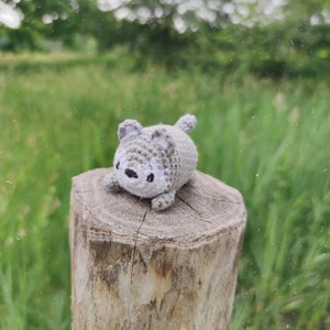 May include: A small gray crocheted wolf plushie with white accents sits on a wooden stump. The wolf has a cute, cartoonish face and is sitting upright.
