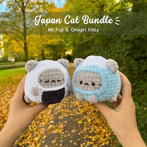May include: Two handmade crochet cat plushies, one white and one light blue, are held up against a blurred background of fall foliage. The white cat is designed as Mt. Fuji, and the blue cat is designed as Onigiri Kitty. The text "Japan Cat Bundle" is at the top.