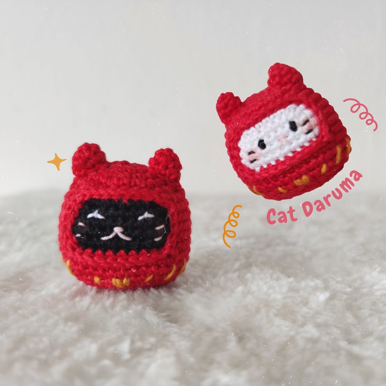 May include: Two red crocheted cat daruma dolls. One is standing upright and the other is floating in the air. Both have black eyes and white whiskers.