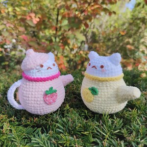 May include: Two crocheted cat plushies, one pink with a strawberry design and the other yellow with a lemon design. Both have white faces with black eyes and pink cheeks.