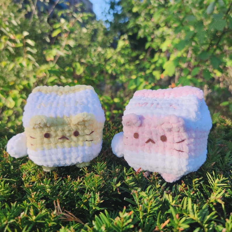 May include: Two crocheted cat-shaped plushies, one yellow and white, the other pink and white, sitting on a bed of green moss.