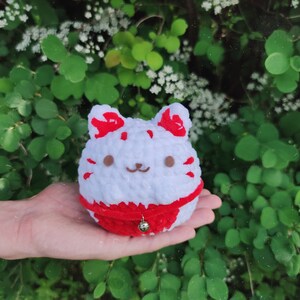 May include: White crocheted cat plush toy with red ears and a red velvet collar with a bell. The toy is sitting in a hand.