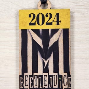 May include: A yellow and black wooden ornament with the text "2024" and "Beetlejuice The Musical" on it.