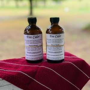May include: Two glass bottles of Fire Cider, a fermented apple cider vinegar drink. The bottles are labeled with the ingredients and instructions for use. The bottles are sitting on a red and white striped fabric.