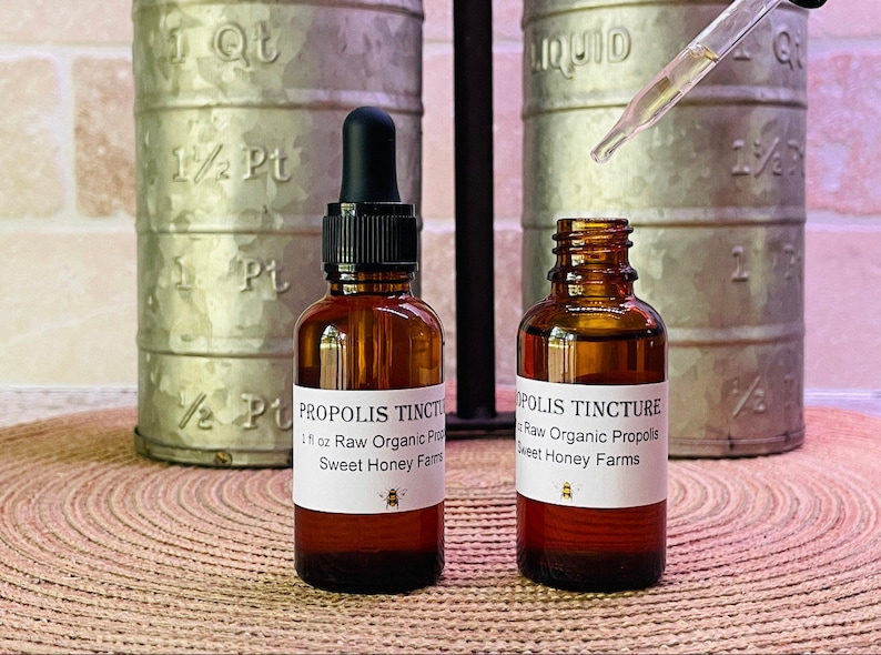 Puede incluir: Dos botellas de vidrio &aacute;mbar con goteros, etiquetadas como "Tintura de Pr&oacute;polis" y que contienen 30 ml de prop&oacute;lis org&aacute;nica cruda. Las botellas est&aacute;n sobre una estera tejida marr&oacute;n, con una taza medidora de metal en el fondo.