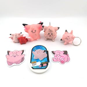 May include: A collection of pink Clefairy items including four small figurines, one keychain, two stickers, and a rectangular card with the text "ピッピ Clefairy" and "80". The items feature the character's signature pink color, pointed ears, and curled hair.