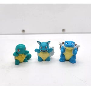 May include: Three small, colourful figurines of Pokémon characters. The figurines are in shades of blue, yellow, and green. The figures are standing on a white surface.