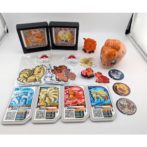 May include: A collection of Pokémon Vulpix and Ninetales merchandise including two framed cards, a small orange figurine, a wrapped orange plush, various stickers with Japanese text, four rectangular game cards, and three circular tokens.