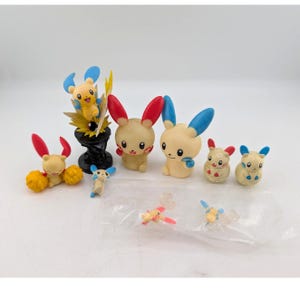 May include: A collection of small, colorful Pikachu figurines. The figures feature various poses and designs, with yellow, red, and blue accents. Some are standing on a black base, while others are in mid-air. The collection includes several different sizes.