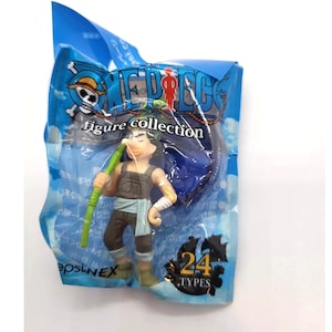 Puede incluir: Un juguete de la colección de figuras de One Piece en un paquete de plástico azul. La figura es de un personaje con una nariz larga, que lleva un atuendo marrón y sostiene un bastón verde. El paquete tiene las palabras "ONE PIECE" y "figure collection".