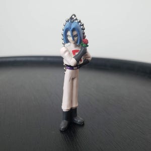 May include: A blue-haired anime character figurine wearing a white shirt with a red emblem and black pants. The figurine is holding a red rose and is attached to a silver chain.