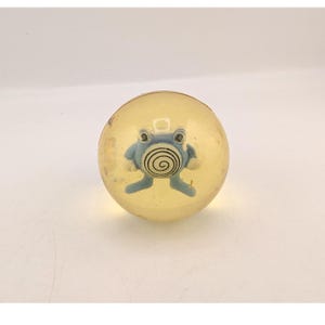 May include: A spherical, translucent yellow bouncy ball containing a small, light blue figure with a prominent black spiral pattern on its belly. The figure has two eyes, two arms, and two legs, appearing suspended within the clear, round object.