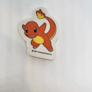 Puede incluir: Gomma de borrar de dibujos animados naranja y amarilla con cola de llama. La goma tiene forma de personaje Pokemon y tiene el texto "©1997 Nintendo/Pokemon."