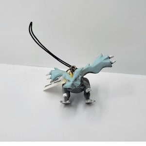 Puede incluir: Pequeña figura gris y azul claro de una criatura con forma de dragón con alas extendidas. La figura tiene una cuerda negra para colgar. Una pequeña etiqueta está adjunta.