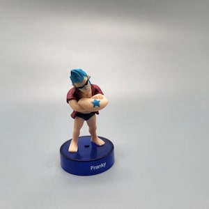 Puede incluir: Figura de un personaje con pelo azul, gafas de sol y camisa roja, de pie sobre una base azul. El personaje sostiene un objeto en forma de estrella. La base está etiquetada como "Franky".