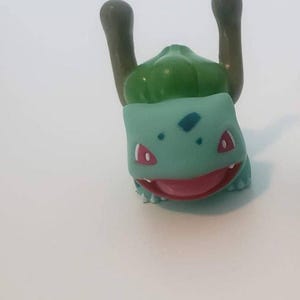 May include: A small, light teal and green plastic figurine of a creature with a large bulbous structure on its back, two brown vine-like appendages extending upward, red eyes, and an open mouth showing a pink interior and small white teeth.