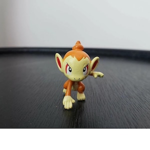 Könnte beinhalten: Kleine, orange und gelbe Figur einer affenähnlichen Kreatur mit roten Akzenten. Die Figur befindet sich in einer hockenden Pose mit einem ausgestreckten Arm. Die Figur hat große Ohren und einen spitzen Kopf. Dunkler Hintergrund.
