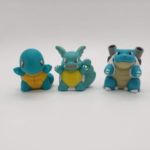 May include: Three blue and yellow cartoon character figurines. The figurines are in different poses and have varying features. The figurines are on a white surface.