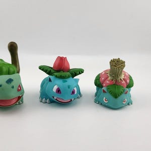 May include: Three Bulbasaur figurines in different poses. The figurines are primarily teal with green and red accents. Each has a different plant-like growth on its back. The figurines are on a white surface.