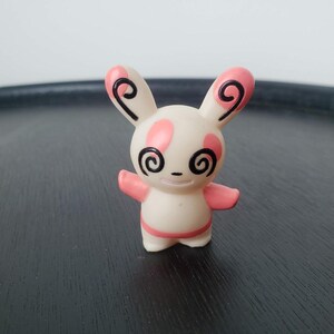 May include: A small, cream-colored figurine of a rabbit-like creature with pink patches on its ears, head, and body. It features black spiral patterns on its ears and eyes, a small horizontal line for a mouth, and two outstretched arms.