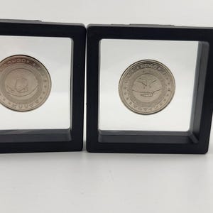 Può includere: Due teche nere quadrate che contengono monete d'argento. Le monete hanno un design circolare con testo e un emblema. Le teche sono su una superficie bianca.