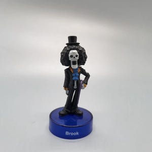 Puede incluir: Figura del personaje Brook de la serie de anime One Piece. La figura es un esqueleto que lleva un sombrero de copa negro, un traje y una corbata azul. La figura se encuentra sobre una base azul con el nombre "Brook" impreso.