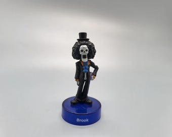 Puede incluir: Figura del personaje Brook de la serie de anime One Piece. La figura es un esqueleto que lleva un sombrero de copa negro, un traje y una corbata azul. La figura se encuentra sobre una base azul con el nombre "Brook" impreso.