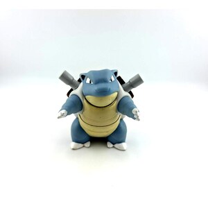 May include: A blue and cream-colored bipedal creature figurine with a rounded head, pointed ears, and two gray cylindrical cannons protruding from its back. The figure has a smooth, segmented shell on its chest and white claws on its feet.