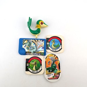 Peut inclure: Un ensemble de cartes à collectionner Pokémon et une petite figurine. Les cartes présentent divers personnages Pokémon, dont une créature verte et jaune en forme de serpent. La figurine est une représentation 3D du même personnage.