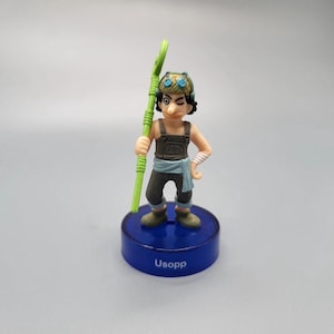 Puede incluir: Una figura del personaje Usopp de la serie de anime One Piece. La figura está sobre una base azul con el nombre "Usopp" impreso. Usopp se representa con un bastón verde, usando un sombrero, un mono y una venda en el brazo.