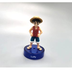 Puede incluir: Figura de un personaje de dibujos animados, con sombrero de paja, chaleco rojo, pantalones cortos azules y sandalias. La figura está sobre una base azul con el nombre "Luffy" impreso. El personaje tiene pelo negro y ojos grandes.