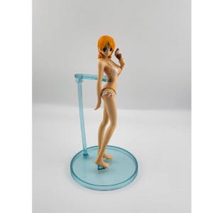 May include: A standing anime-style figurine with short orange hair, wearing a light pink bikini, and holding a small brown ice cream cone. The figure is supported by a translucent blue plastic stand consisting of a circular base and a vertical arm.