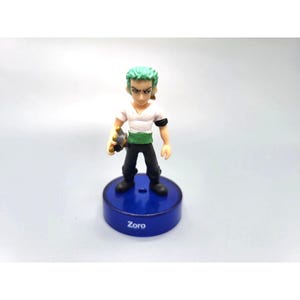 Puede incluir: Figura del personaje Zoro de la serie de anime One Piece. La figura tiene el pelo verde, una camisa blanca, pantalones negros y una faja verde. Se encuentra sobre una base azul con el nombre "Zoro" impreso.