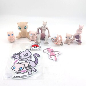 May include: A collection of Pokemon figurines and stickers. The figurines are in various poses and colors, including pink, purple, and tan. The stickers feature Mew and Mewtwo characters. A Pokemon logo sticker is also included.