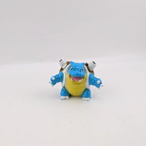 May include: A small blue and yellow creature figurine with a brown shell on its back. The figure has two white cannons protruding from the shell, white claws on its hands, and an open mouth showing a pink tongue and white teeth.